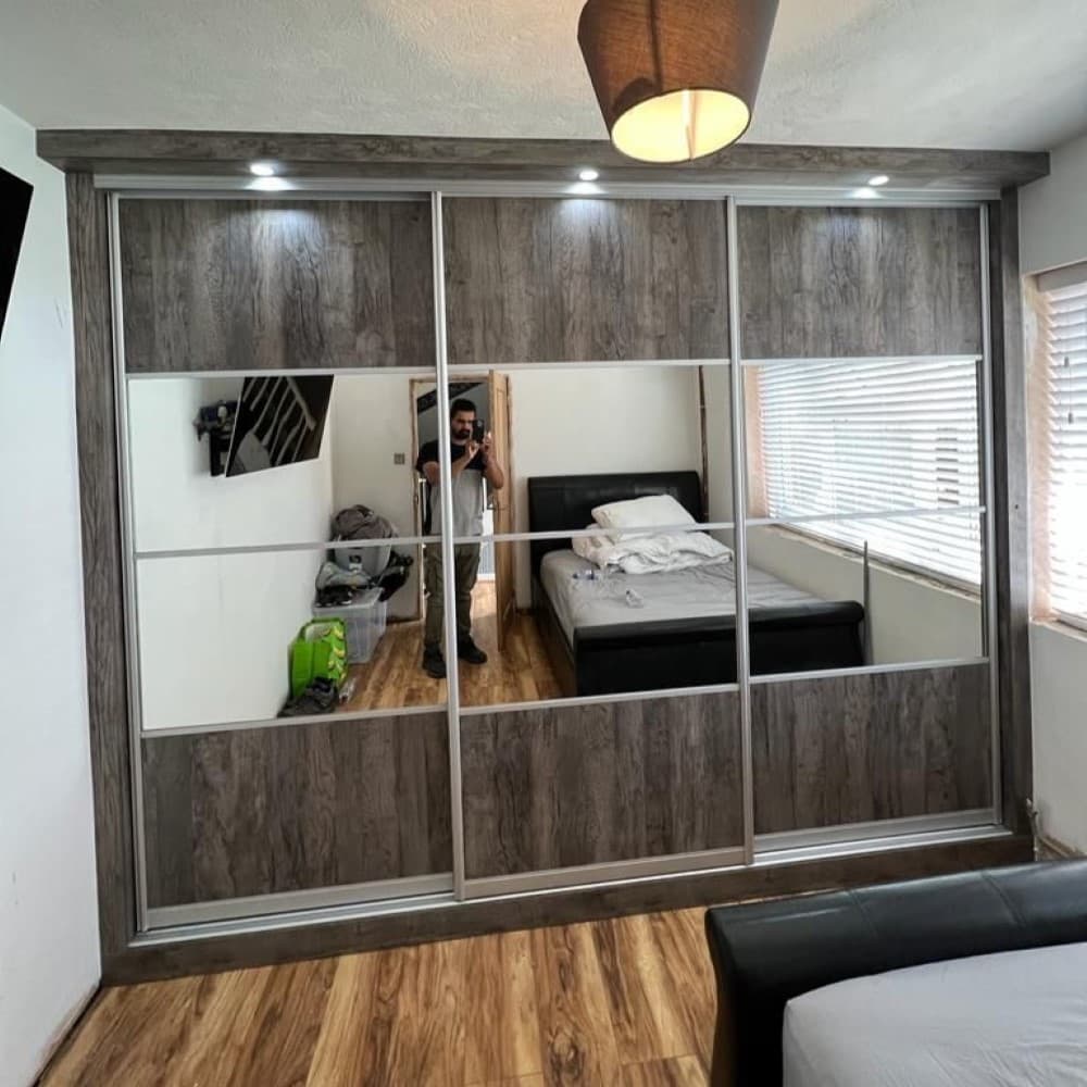 Woodgrain effect sliding door wardrobe in grey tones with overhead LED lighting and mirrored panels in a fitted bedroom.